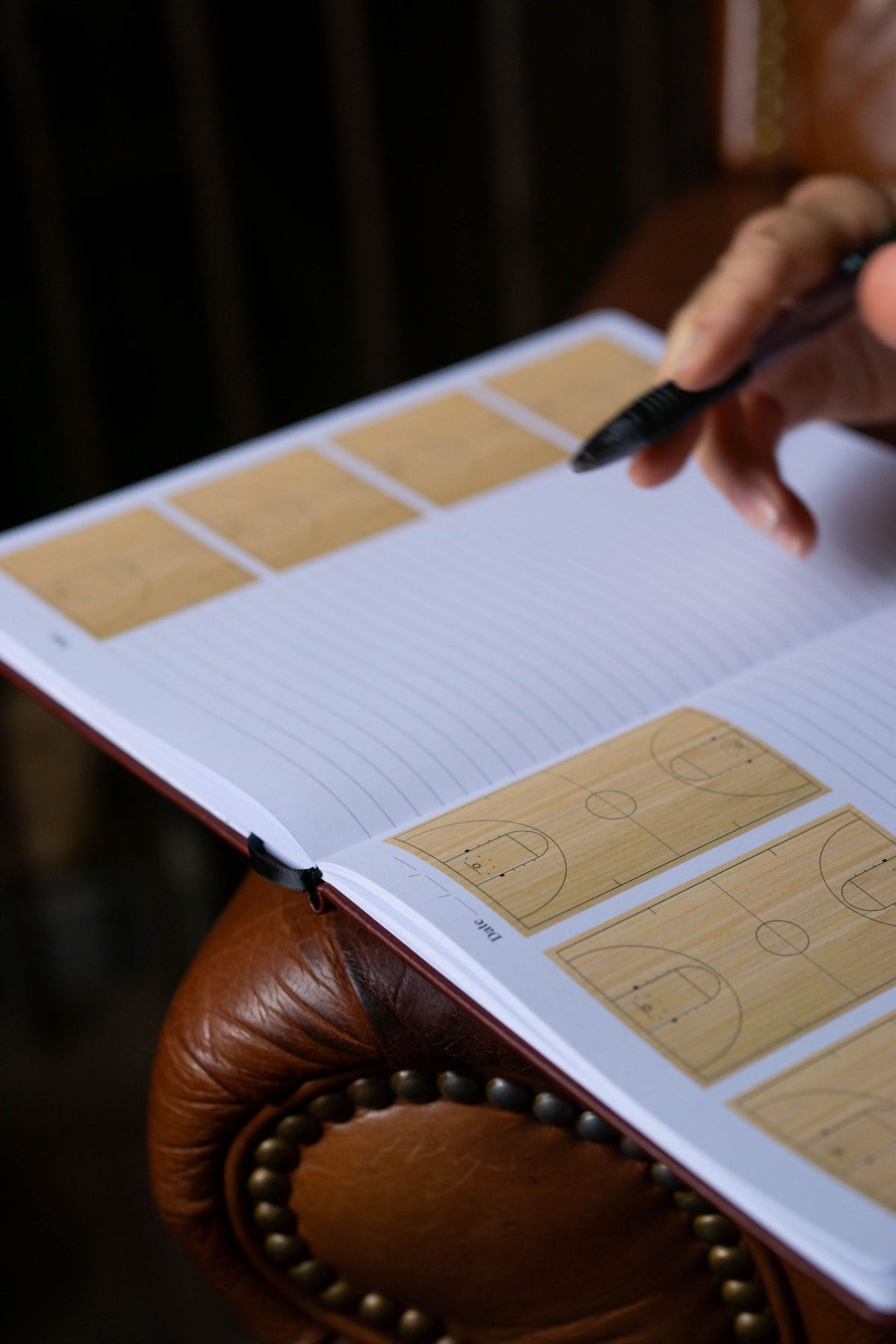 A hand holding a pen over the Basketball Edition journal, open to basketball court diagrams and lined pages