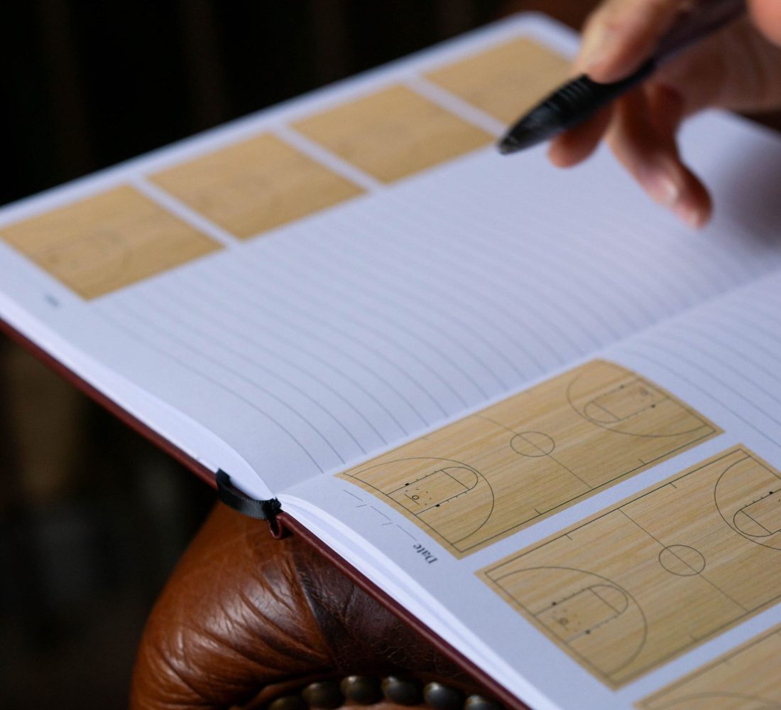 A coach's hand with a pen over the open journal showing basketball court diagrams and lined pages